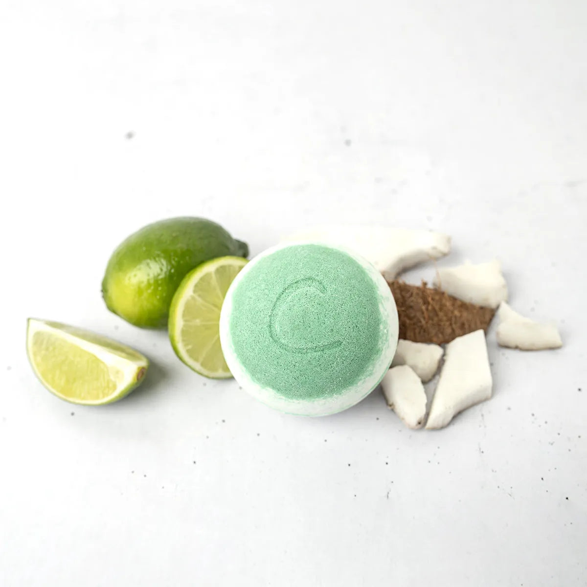 Round light green bath bomb with a letter C, surrounded by fresh lime wedges and coconut chunks on a white surface.
